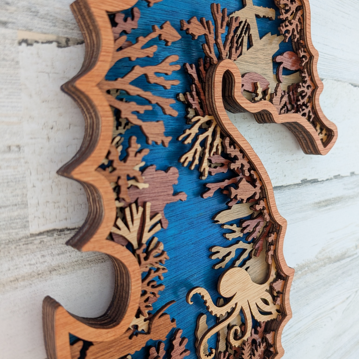 Laser-cut wooden seahorse wall art with layered wood tones and an underwater scene featuring coral, fish, and an octopus on a blue background, mounted on a white distressed wood surface.