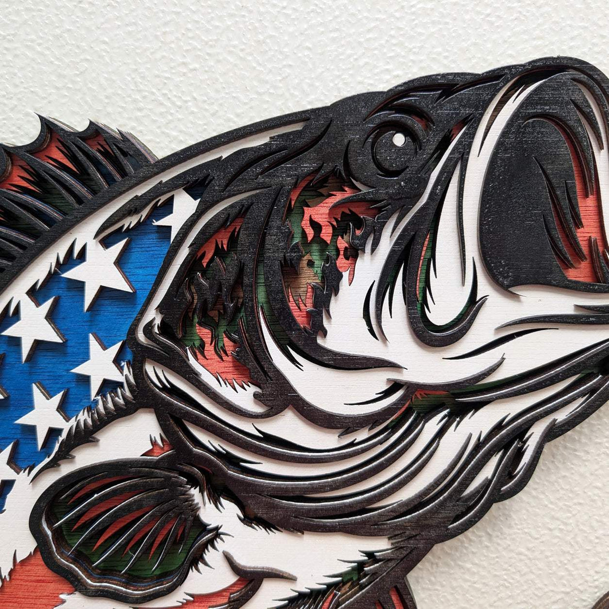 A close-up of a layered wooden wall art piece depicting a largemouth bass fish in profile, with cutout layers showing red, green, and blue wood tones over a white background with white stars.