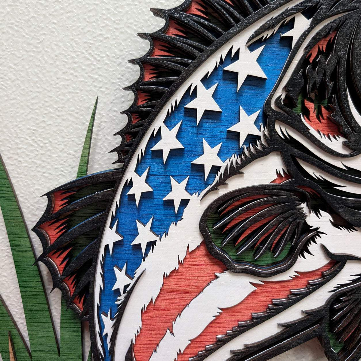 Close-up of a layered wooden wall art piece featuring a stylized fish design with white stars on a blue field and red-and-white striped accents, mounted against a light textured wall.