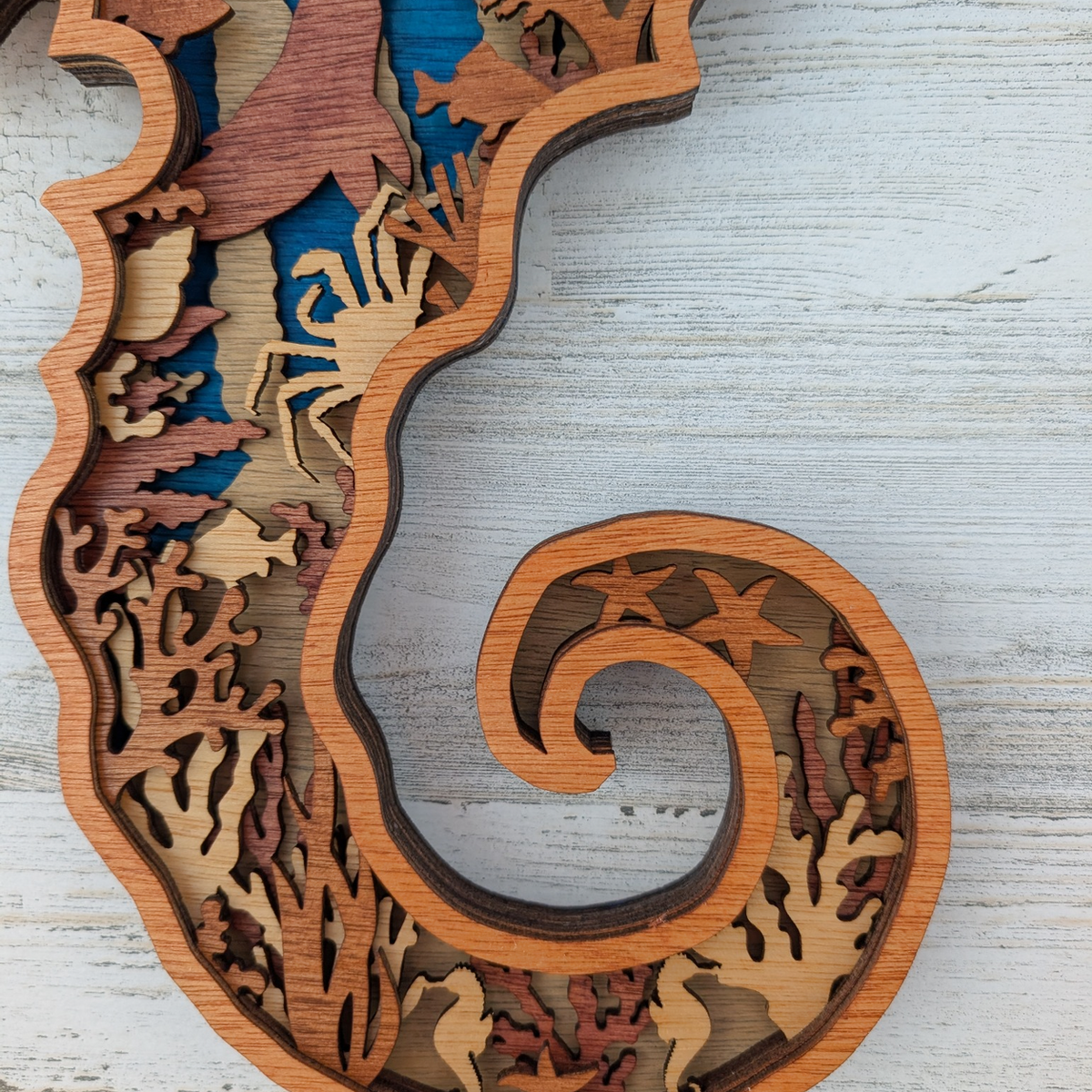 A laser-cut wooden seahorse wall decor piece with layered wood tones and cutout details, photographed on a light weathered wood surface.
