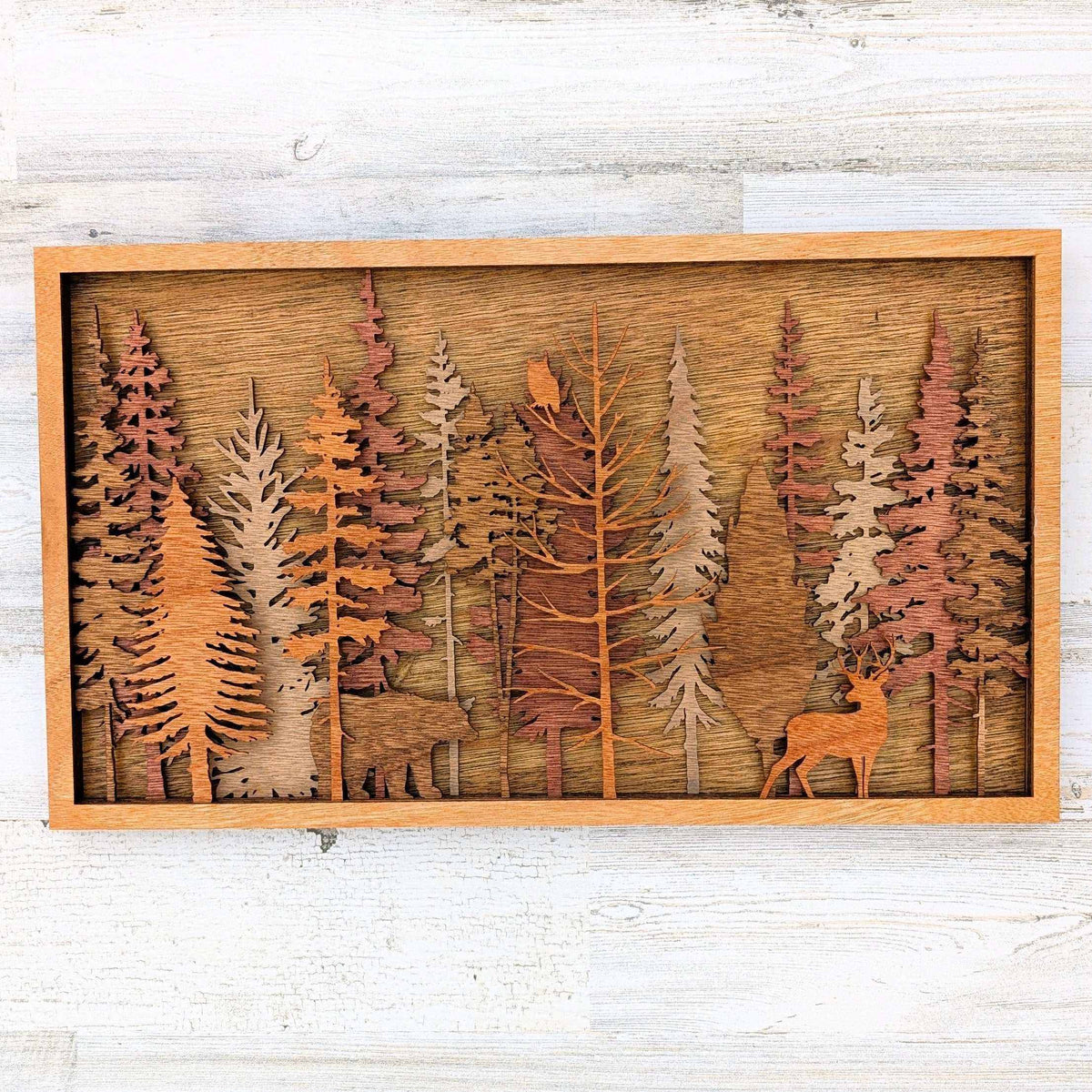 Handcrafted wood art featuring a layered forest scene with colorful evergreen trees and a carved buck in warm wood tones